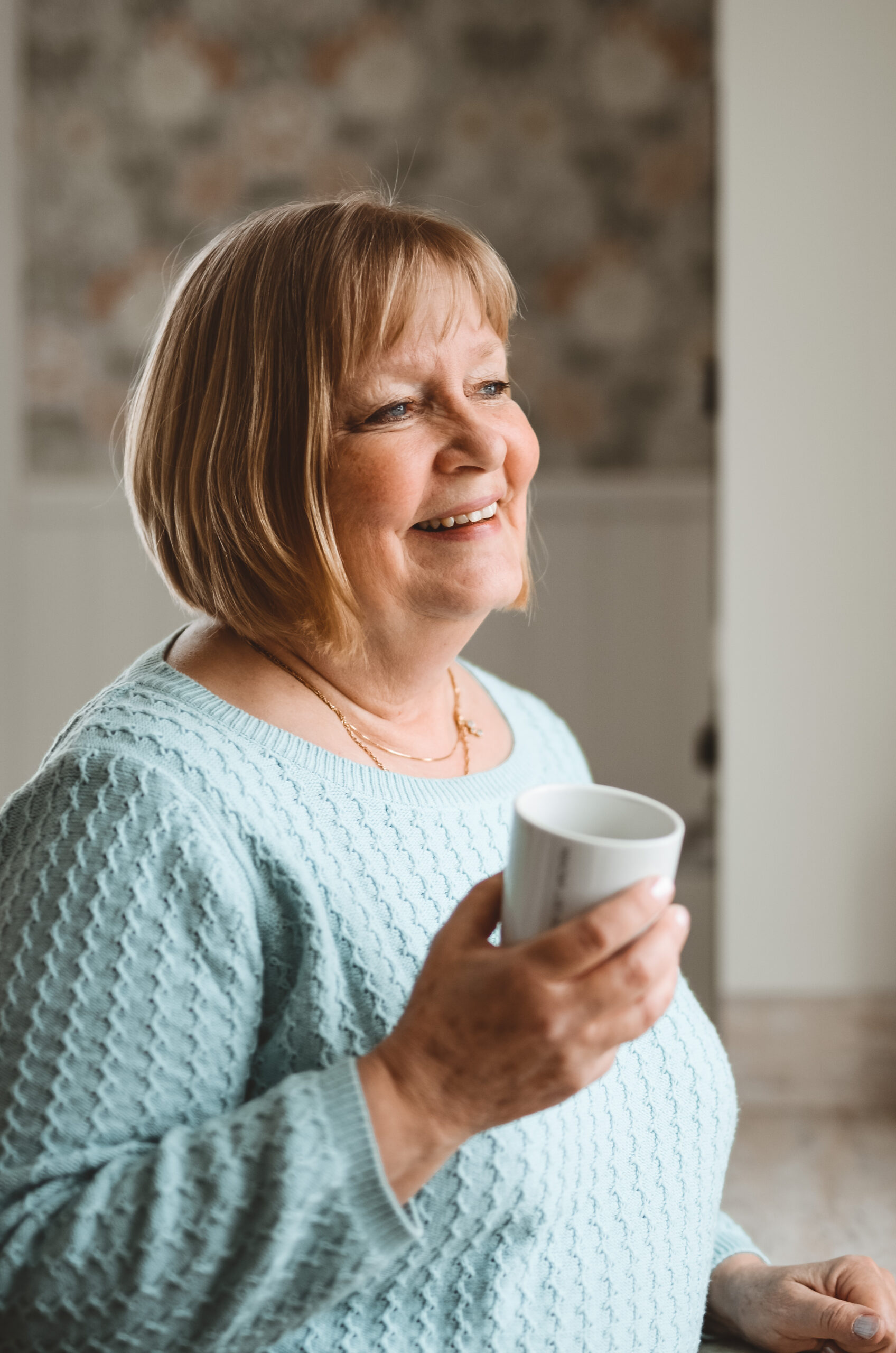 A portrait of Mamma Pia in her kitchen, in a turquoise knitted sweater and a blonde page, holding a cup of coffee in her hand.